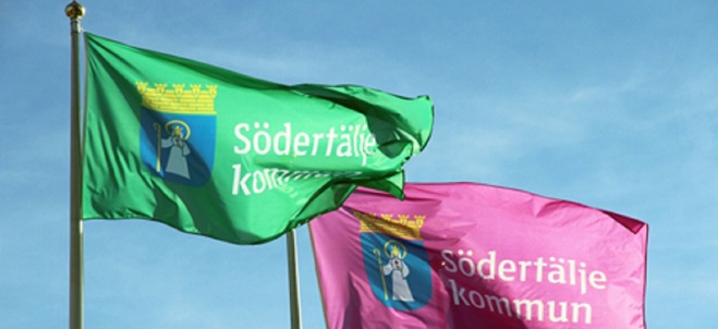 Bild på två av Södertälje kommuns flaggor. Flaggorna är hissade på hel stång och på flaggorna står kommunens logotyp bestående av Sankta Ragnhild samt ordbilden Södertälje kommun.