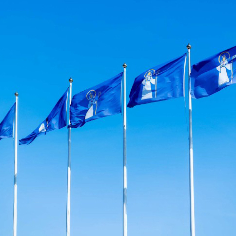 Bilden visar Södertälje kommun-flaggor som vajar i vinden mot en blå himmel.