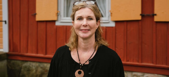 Eva Skyllberg i svart klänning och glasögon på huvudet framför en röd och gul äldre byggnad på Torekällberget