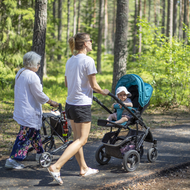 Bild på två personer som promenerar längs en gångväg i skogen. En med barnvagn och en med rullator.