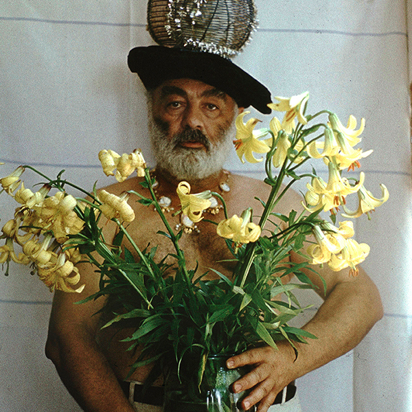 Sergej Parajanov står med bar överkropp och hatt. Han håller i en vas med gula blommor.