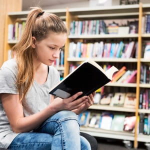 Flicka läser bok i bibliotek.