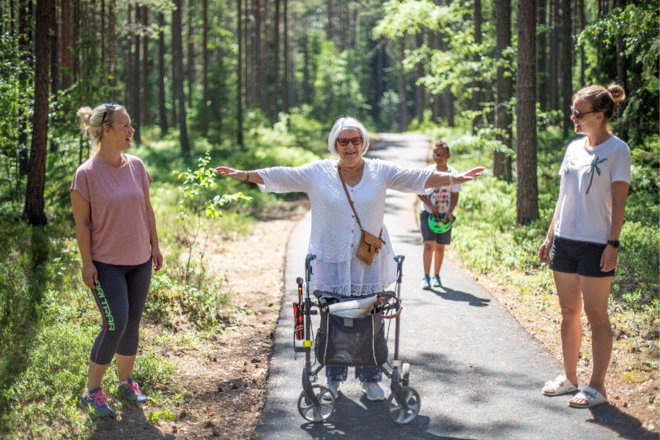 Tre kvinnor i olika åldrar står på en gångväg i skogen. Kvinnan i mitten står bakom en rullator och sträcker ut armarna åt sidorna. . Solen skiner på dem. I bakgrunden syns ett barn i tioårsåldern som håller en cykelhjälm i händerna. 