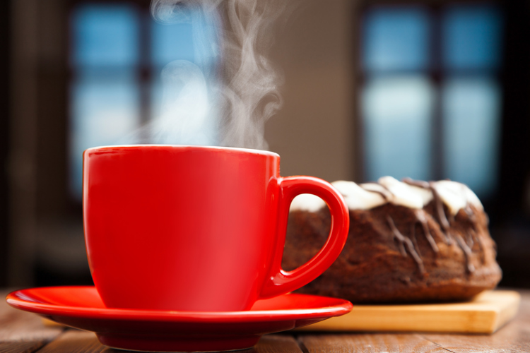 Bild på en röd mugg på fat med rykande kaffe och en sockerkaka i bakgrunden.