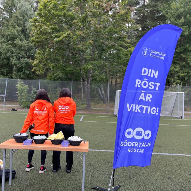 Bilden visar två personer som står med ryggen mot kameran. På deras tröjor står det Södertälje röstar och valinformatör. Bredvid står en beachflagga där det står "Din röst är viktig".