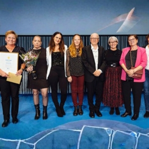 Nio personer står på rad på en scen. De håller i blommor och diplom.