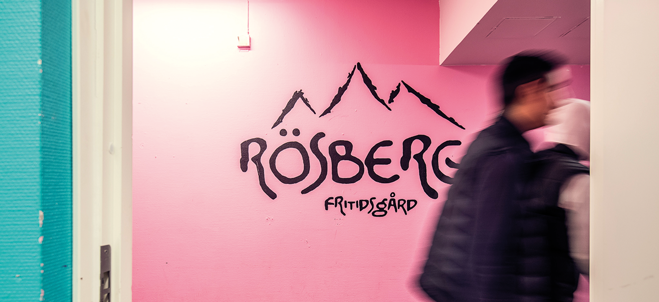 Det står Rösberga fritidsgård i svart på en rosa vägg. Två ungdomar syns.