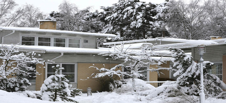 En byggnad med snö på taket och på gårdsplanen. Träd och buskar har snö på sina grenar.