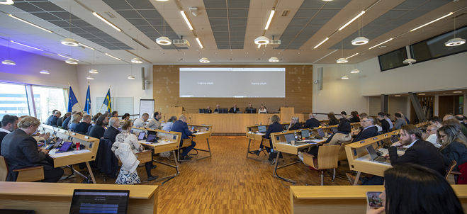 Bilden visar ett kommunfullmäktigemöte. I bilden finns flertalet deltagande som sitter vid bord som är riktade mot en scen längre fram i rummet.