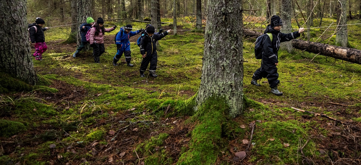 En grupp barn följer en stig i skogen hållandes i ett rep.